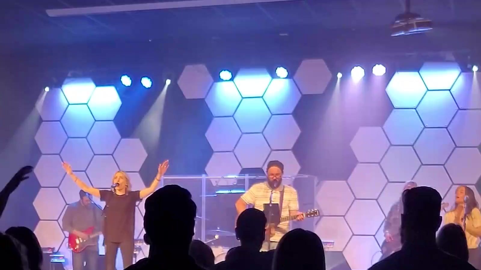 A worship band on a lit stage with people raising their hands